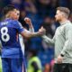 Chelsea midfielder Enzo Fernández (wearing the blue number 8 jersey) shakes hands and converses with interim head coach Calum McFarlane (in a grey Chelsea training hoodie) on a football pitch during a match day. Image used for Sportxparte news.