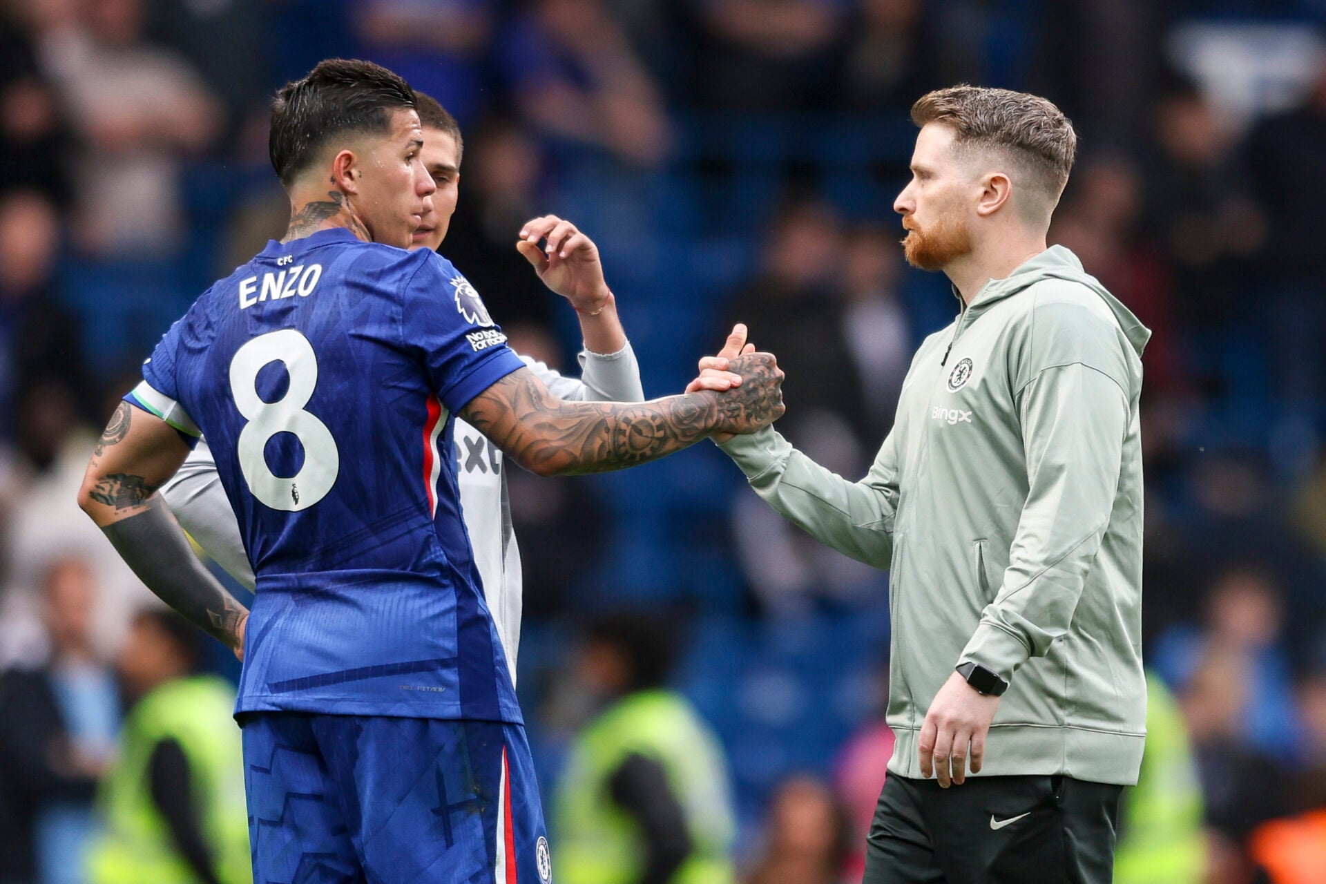 Chelsea midfielder Enzo Fernández (wearing the blue number 8 jersey) shakes hands and converses with interim head coach Calum McFarlane (in a grey Chelsea training hoodie) on a football pitch during a match day. Image used for Sportxparte news.