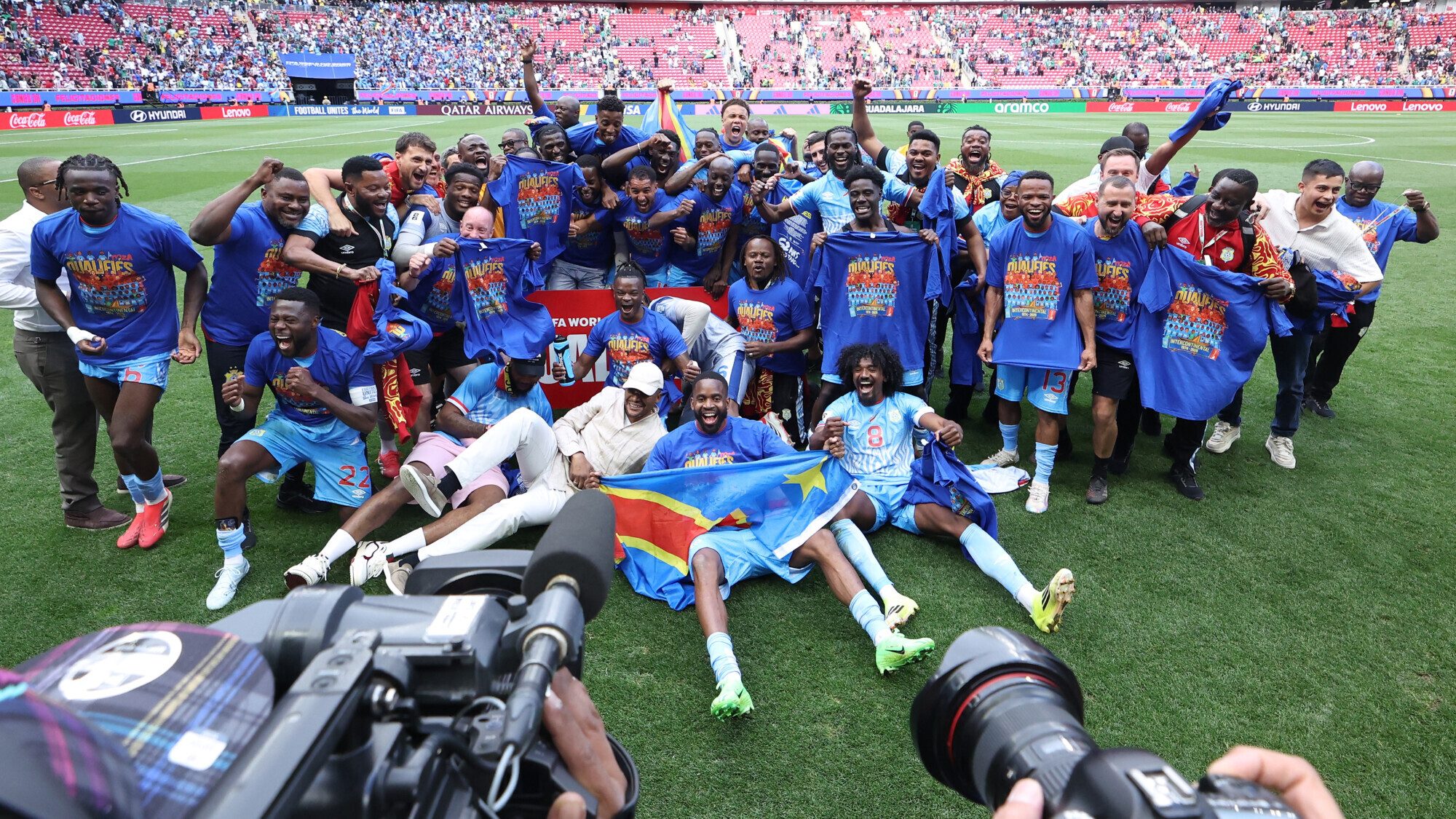 DR Congo national football team celebrating on a vibrant green pitch after a match. Dozens of players, coaching staff, and team officials in matching bright blue jerseys and training gear jump, cheer, and embrace with joyous expressions. Near the center, a player sits on the grass holding up the blue, red, and yellow flag of the Democratic Republic of the Congo. Image used for Sportxparte news.