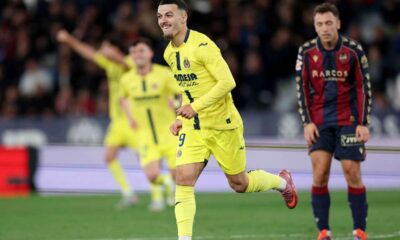 Georges Mikautadze is celebrating a goal for Villarreal CF. He is wearing a bright yellow kit and is captured mid-run with a joyful expression and clenched fists. In the background, teammates in yellow and an opposing player in a navy and maroon kit look on. Image used for Sportxparte Preview.