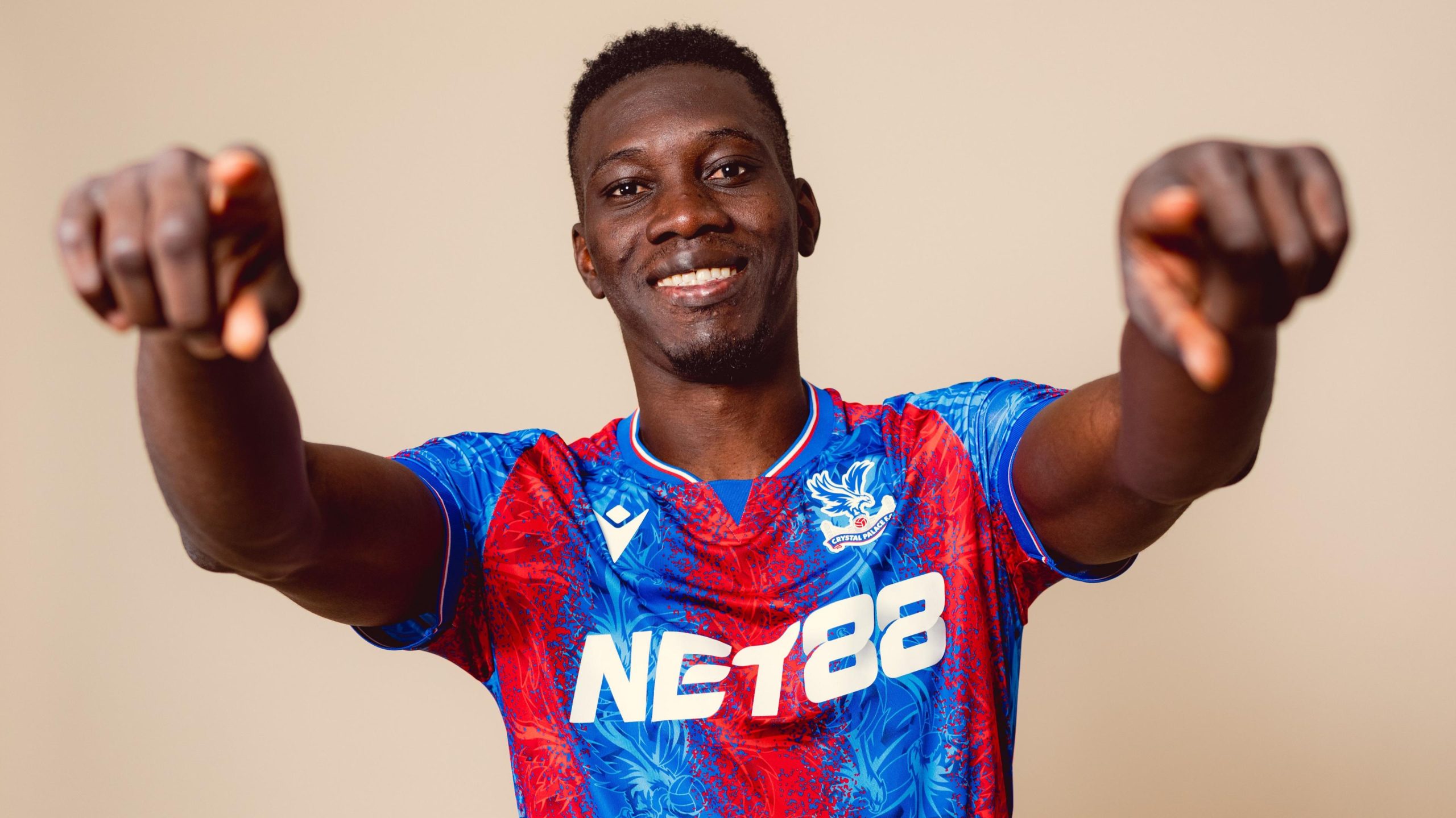 A smiling, medium shot of Senegalese football player Ismaïla Sarr against a plain, neutral background. He is wearing the red and blue Crystal Palace 2024/25 home jersey with the "NET88" sponsor logo clearly visible across the front. Image used for Sportxparte news. 