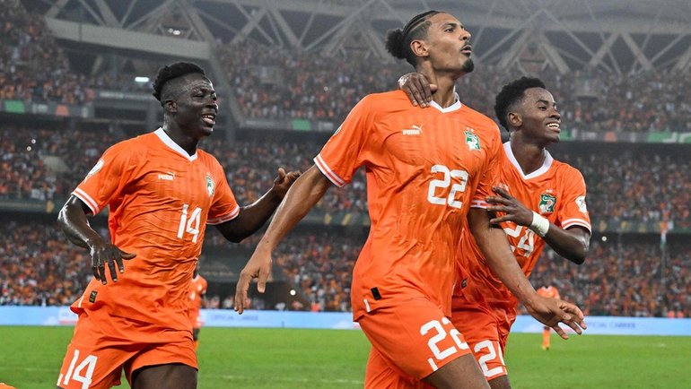 Ivory Coast national football team celebrate joyfully on the pitch during the 2023 Africa Cup of Nations (AFCON) final. Center-forward Sébastien Haller, wearing the bright orange number 22 jersey, smiles broadly with his arms slightly raised as a teammate hugs him from behind. To his right, Oumar Diakité, wearing jersey number 14, joins the celebration with an open-mouthed expression of excitement. Image used for Sportxparte news.