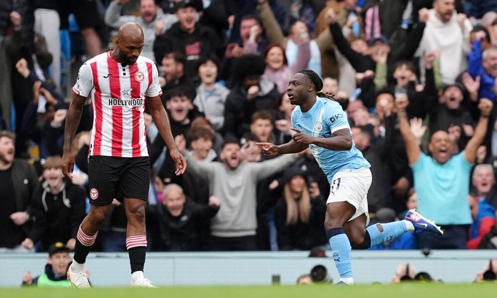 Manchester City winger Jérémy Doku celebrates with a pointed finger and a wide smile after scoring the opening goal in a 3-0 victory over Brentford on May 9, 2026. Beside him, a Brentford player in a red and white striped jersey, Igor Thiago, looks on as the Etihad Stadium crowd cheers in the background. Image used for Sportxparte news roundup.