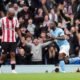 Manchester City winger Jérémy Doku celebrates with a pointed finger and a wide smile after scoring the opening goal in a 3-0 victory over Brentford on May 9, 2026. Beside him, a Brentford player in a red and white striped jersey, Igor Thiago, looks on as the Etihad Stadium crowd cheers in the background. Image used for Sportxparte news roundup.
