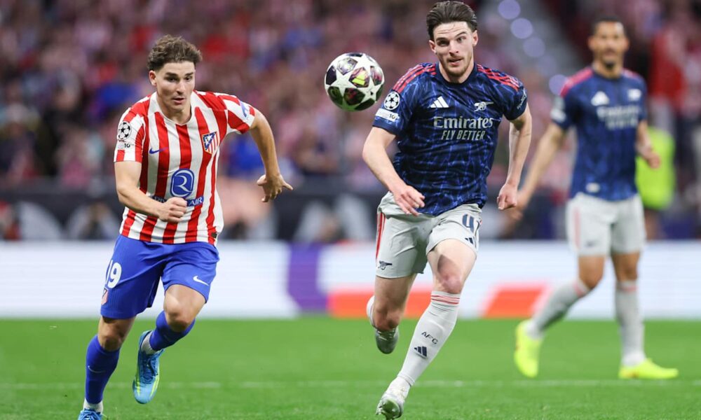 An action shot from a football match featuring Declan Rice of Arsenal and Julián Álvarez of Atlético Madrid competing for the ball. Rice, on the right, is wearing a dark blue and red Arsenal away kit, while Álvarez, on the left, is in Atlético’s traditional red-and-white striped home jersey. Image used for Sportxparte news Preview.