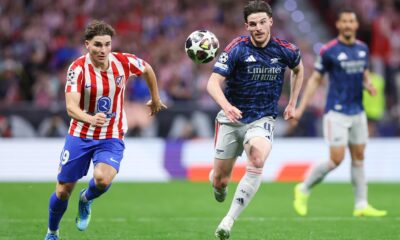 An action shot from a football match featuring Declan Rice of Arsenal and Julián Álvarez of Atlético Madrid competing for the ball. Rice, on the right, is wearing a dark blue and red Arsenal away kit, while Álvarez, on the left, is in Atlético’s traditional red-and-white striped home jersey. Image used for Sportxparte news Preview.