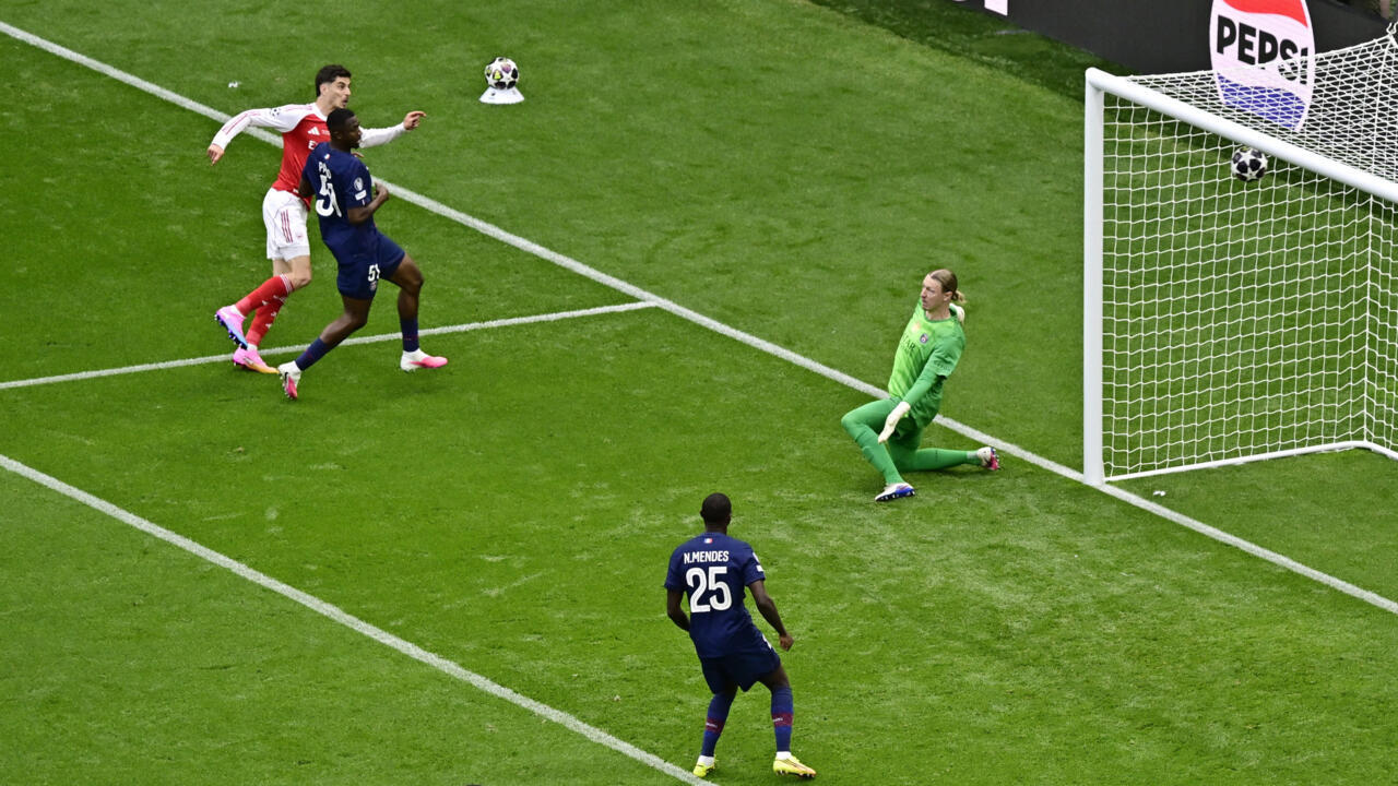 Arsenal forward Kai Havertz is shooting a soccer ball from a tight angle on the left side of the penalty box during the 2026 UEFA Champions League final. Havertz, wearing Arsenal’s red and white kit, strikes the ball past a lunging Paris Saint-Germain defender wearing a dark blue kit. Image used for Sportxparte news.