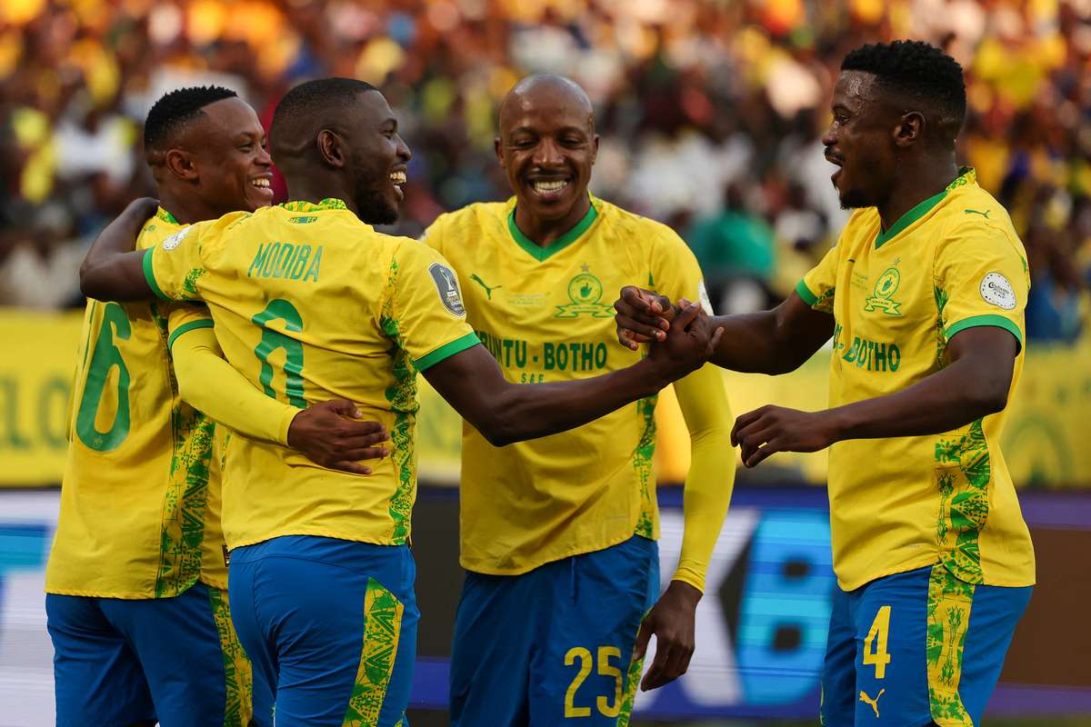 Four Mamelodi Sundowns football players, including Aubrey Modiba and Teboho Mokoena, celebrate a goal on the pitch wearing the yellow and blue home kit. Image used for Sportxparte news. 