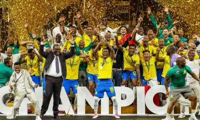 A jubilant squad of Mamelodi Sundowns football players in yellow and blue uniforms celebrates on a stage covered in gold confetti. A player in the center holds up the gold CAF Champions League trophy. Image used for Sportxparte news.