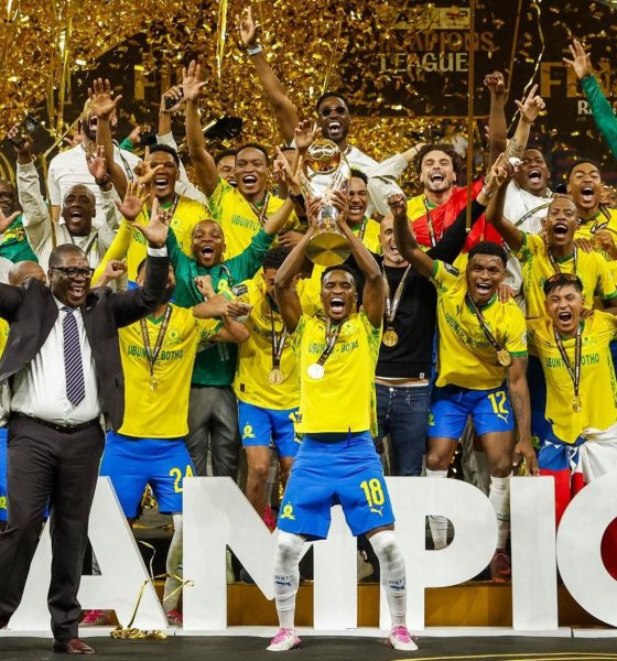 A jubilant squad of Mamelodi Sundowns football players in yellow and blue uniforms celebrates on a stage covered in gold confetti. A player in the center holds up the gold CAF Champions League trophy. Image used for Sportxparte news.