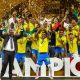 A jubilant squad of Mamelodi Sundowns football players in yellow and blue uniforms celebrates on a stage covered in gold confetti. A player in the center holds up the gold CAF Champions League trophy. Image used for Sportxparte news.