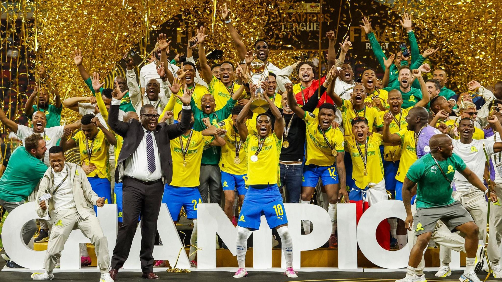 A jubilant squad of Mamelodi Sundowns football players in yellow and blue uniforms celebrates on a stage covered in gold confetti. A player in the center holds up the gold CAF Champions League trophy. Image used for Sportxparte news.