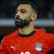 Mohamed Salah representing the Egyptian National Football Team. He is seen sporting a shorter hairstyle and trimmed beard while wearing the official Egyptian red Puma national jersey. Image used for Sportxparte news.
