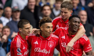 An image of four professional football players from Nottingham Forest celebrating a goal. They are wearing their red home kits with white pinstripes and "Bally's" sponsorship across the chest. From left to right, Nicolas Domínguez, Igor Jesus, James McAtee (background), and Taiwo Awoniyi are huddled together, smiling and embracing in a moment of triumph on the pitch, with a blurred crowd of fans in the background. Image used for Sportxparte news roundup.