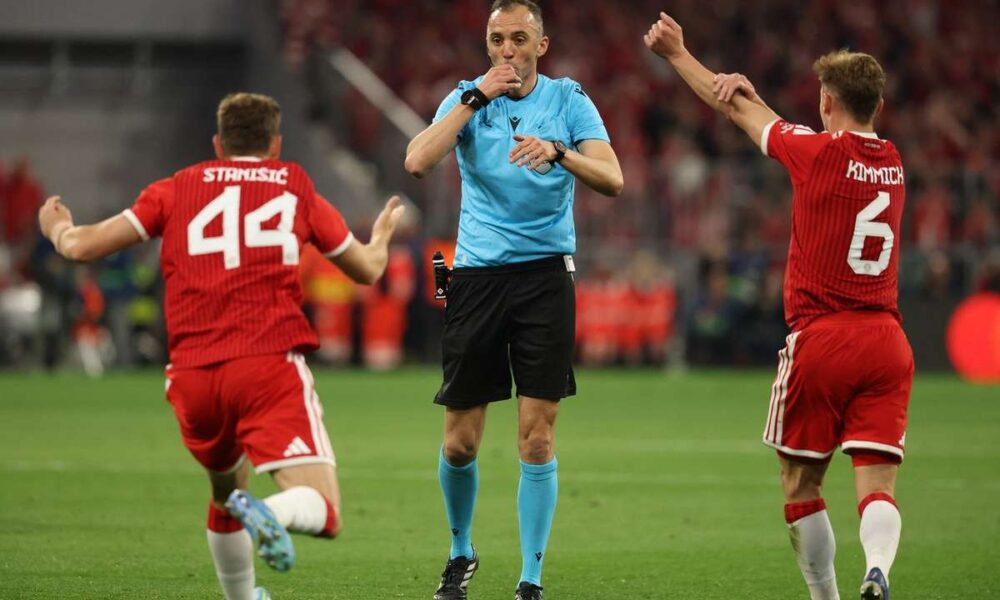 Portuguese referee João Pinheiro in a bright blue uniform, blowing his whistle and pointing. He is flanked by two Bayern Munich players in red jerseys, Josip Stanišić (#44) and Joshua Kimmich (#6), who have their arms raised in protest or appeal against a decision. Image used for Sportxparte news.