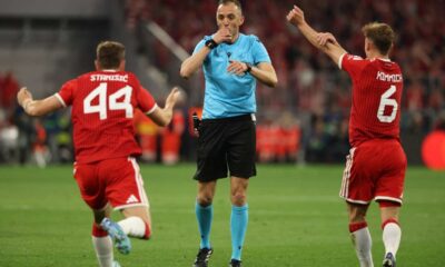 Portuguese referee João Pinheiro in a bright blue uniform, blowing his whistle and pointing. He is flanked by two Bayern Munich players in red jerseys, Josip Stanišić (#44) and Joshua Kimmich (#6), who have their arms raised in protest or appeal against a decision. Image used for Sportxparte news.