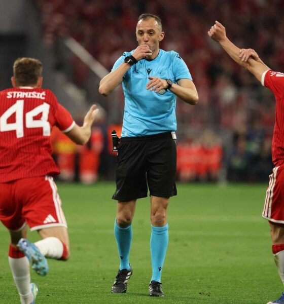 Portuguese referee João Pinheiro in a bright blue uniform, blowing his whistle and pointing. He is flanked by two Bayern Munich players in red jerseys, Josip Stanišić (#44) and Joshua Kimmich (#6), who have their arms raised in protest or appeal against a decision. Image used for Sportxparte news.