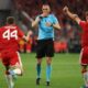 Portuguese referee João Pinheiro in a bright blue uniform, blowing his whistle and pointing. He is flanked by two Bayern Munich players in red jerseys, Josip Stanišić (#44) and Joshua Kimmich (#6), who have their arms raised in protest or appeal against a decision. Image used for Sportxparte news.