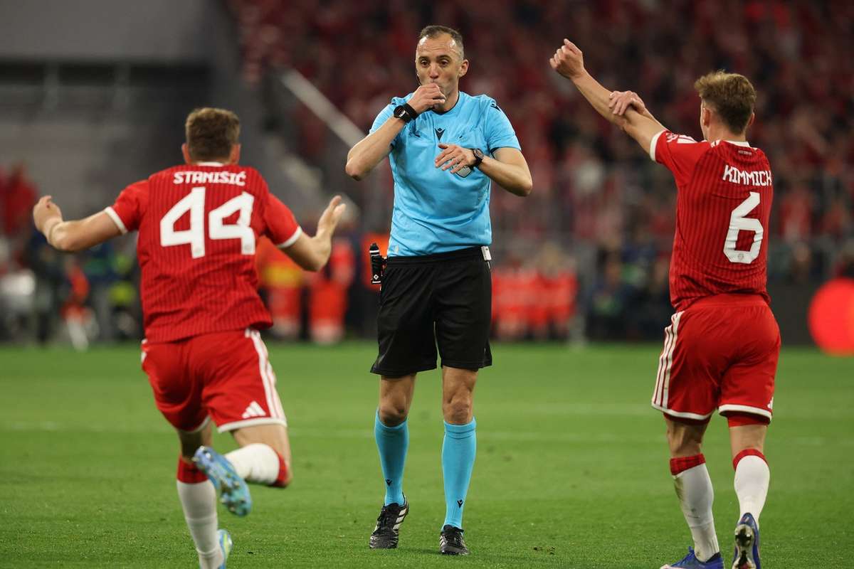 Portuguese referee João Pinheiro in a bright blue uniform, blowing his whistle and pointing. He is flanked by two Bayern Munich players in red jerseys, Josip Stanišić (#44) and Joshua Kimmich (#6), who have their arms raised in protest or appeal against a decision. Image used for Sportxparte news.