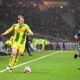 The player in the yellow and green kit is Nicolas Cozza of FC Nantes. The player in the blue kit is Mason Greenwood of Olympique de Marseille, wearing his characteristic number 10. Image used for Sportxparte preview news.