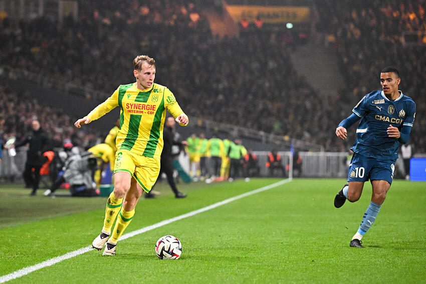 The player in the yellow and green kit is Nicolas Cozza of FC Nantes. The player in the blue kit is Mason Greenwood of Olympique de Marseille, wearing his characteristic number 10. Image used for Sportxparte preview news.