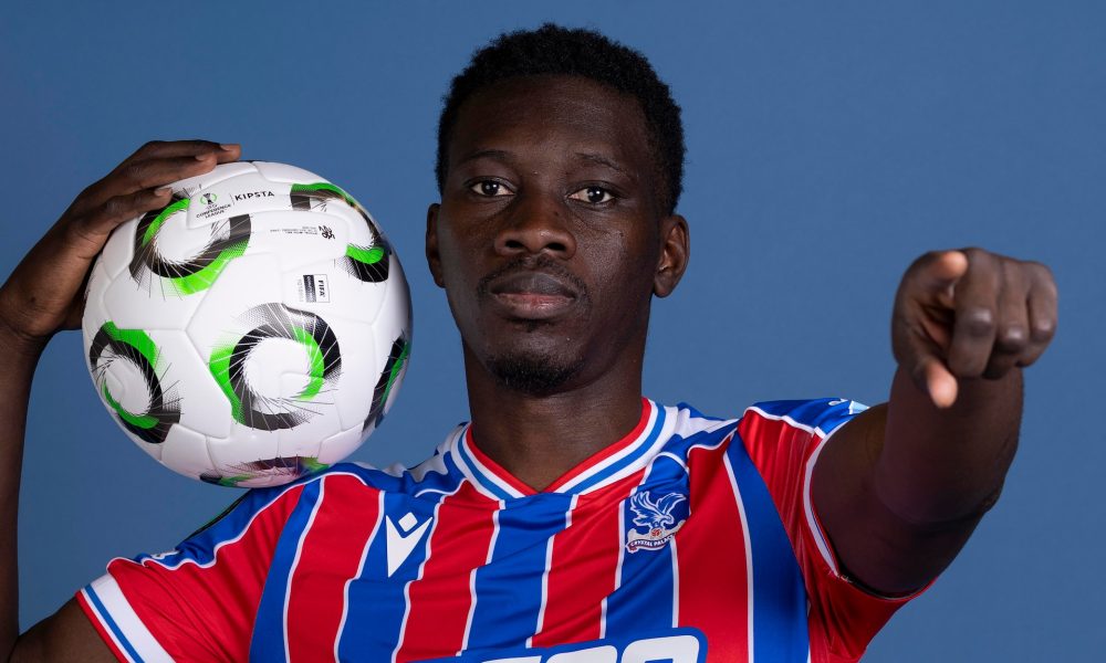 A professional studio portrait of Senegalese professional footballer Ismaïla Sarr, dressed in the red and blue striped Crystal Palace home kit with white trim. He is holding a white Kipsta football with black and green circular patterns on his right shoulder, while extending his left hand to point directly forward. Image used for Sportxparte news.