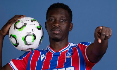 A professional studio portrait of Senegalese professional footballer Ismaïla Sarr, dressed in the red and blue striped Crystal Palace home kit with white trim. He is holding a white Kipsta football with black and green circular patterns on his right shoulder, while extending his left hand to point directly forward. Image used for Sportxparte news.