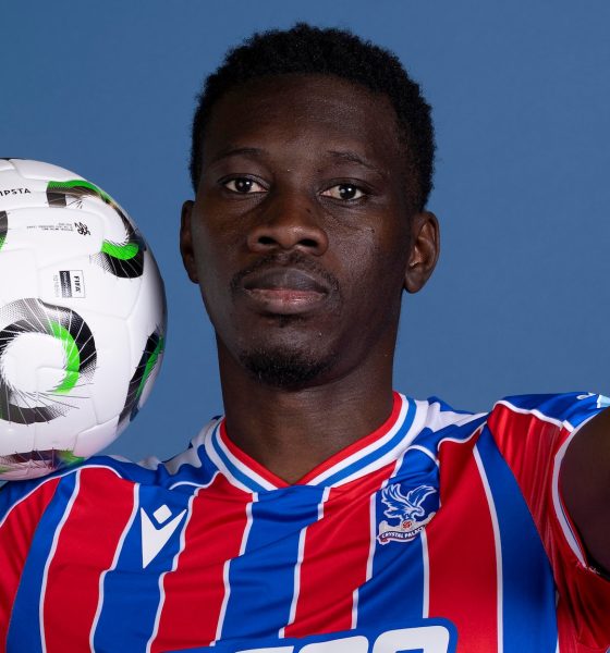 A professional studio portrait of Senegalese professional footballer Ismaïla Sarr, dressed in the red and blue striped Crystal Palace home kit with white trim. He is holding a white Kipsta football with black and green circular patterns on his right shoulder, while extending his left hand to point directly forward. Image used for Sportxparte news.