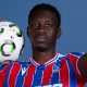A professional studio portrait of Senegalese professional footballer Ismaïla Sarr, dressed in the red and blue striped Crystal Palace home kit with white trim. He is holding a white Kipsta football with black and green circular patterns on his right shoulder, while extending his left hand to point directly forward. Image used for Sportxparte news.