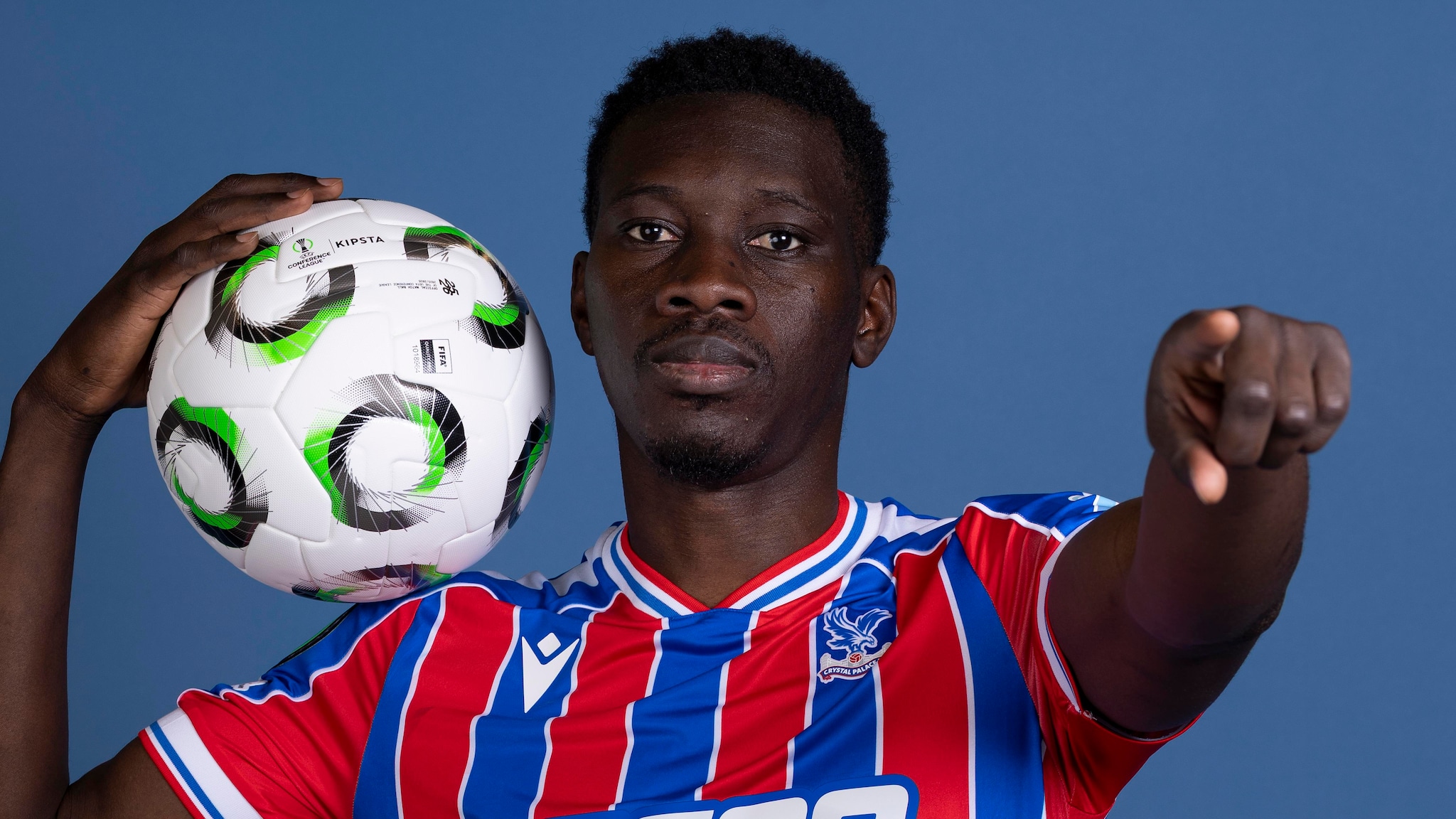 A professional studio portrait of Senegalese professional footballer Ismaïla Sarr, dressed in the red and blue striped Crystal Palace home kit with white trim. He is holding a white Kipsta football with black and green circular patterns on his right shoulder, while extending his left hand to point directly forward. Image used for Sportxparte news.