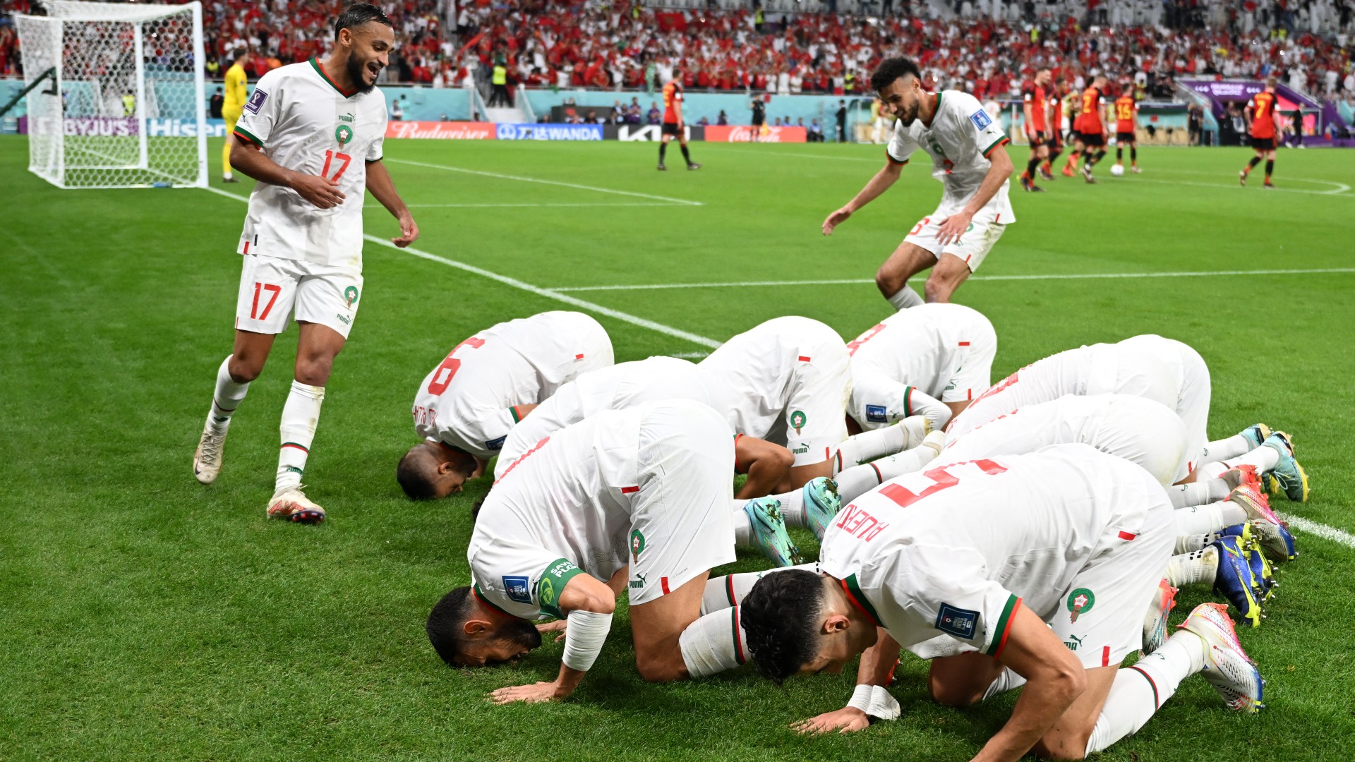 The Morocco national football team is performing the Islamic prostration of gratitude, known as Sujud al-Shukr, to celebrate a historic victory. Standing on the left with jersey number 17 is winger Sofiane Boufal, while the players on the ground express their thanks after defeating Belgium 2-0 in Group F during the 2022 FIFA World Cup at the Al Thumama Stadium. Image used for Sportxparte news.