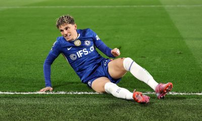 Alejandro Garnacho reacts with a frustrated expression while sitting on the green grass during a match. He wears the blue Chelsea FC home kit with the IFS.ai sponsor logo, white socks, and pink boots. Image used for Sportxparte news.