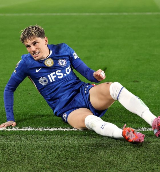 Alejandro Garnacho reacts with a frustrated expression while sitting on the green grass during a match. He wears the blue Chelsea FC home kit with the IFS.ai sponsor logo, white socks, and pink boots. Image used for Sportxparte news.
