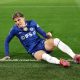 Alejandro Garnacho reacts with a frustrated expression while sitting on the green grass during a match. He wears the blue Chelsea FC home kit with the IFS.ai sponsor logo, white socks, and pink boots. Image used for Sportxparte news.