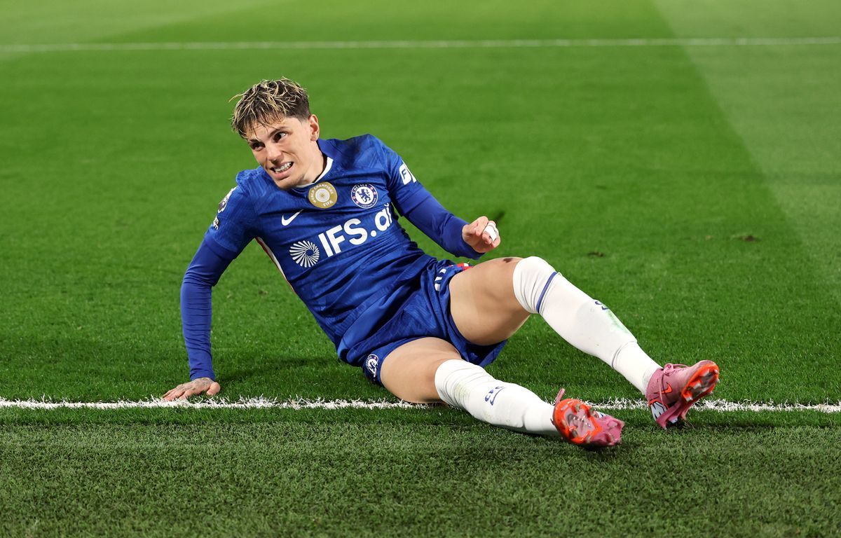 Alejandro Garnacho reacts with a frustrated expression while sitting on the green grass during a match. He wears the blue Chelsea FC home kit with the IFS.ai sponsor logo, white socks, and pink boots. Image used for Sportxparte news.