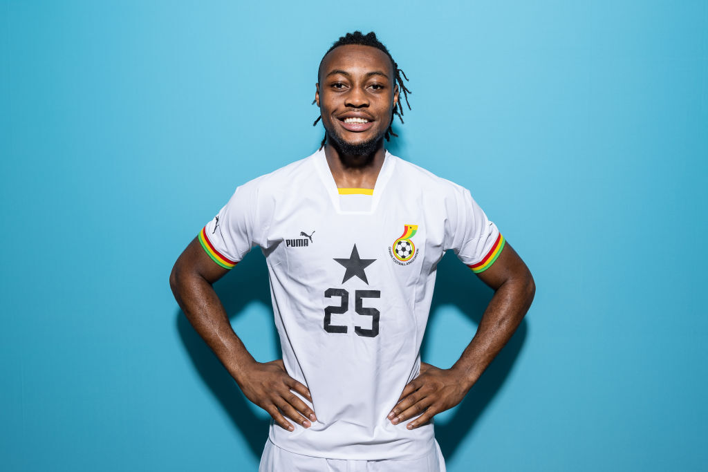 A studio portrait of professional footballer Antoine Semenyo posing against a solid light-blue background. He is smiling warmly at the camera with his hands placed on his hips. He is wearing the white Puma Ghana national football team home jersey, which features a prominent black star and the number 25 printed on the center of the chest, alongside the Ghana Football Association badge. Image used for Sportxparte news.