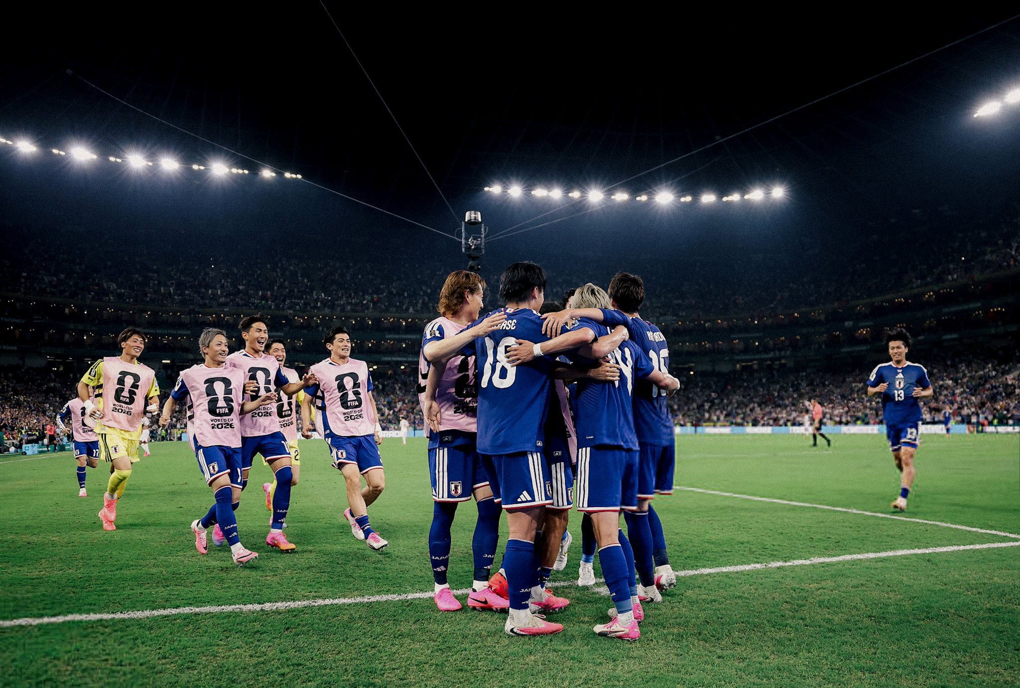 Japan national football team players celebrating a goal in blue Adidas jerseys and pink FIFA World Cup 2026 warmup bibs on a stadium pitch under bright lights. Image used for Sportxparte news.