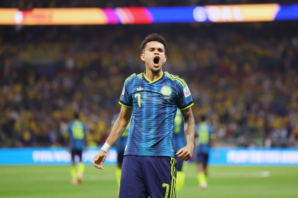 Colombian footballer Luis Díaz celebrating passionately with mouth wide open after scoring a goal for Colombia in the 2024 Adidas dark blue away jersey with neon green accents at the 2026 FIFA World Cup stadium match. Image used for Sportxparte news.