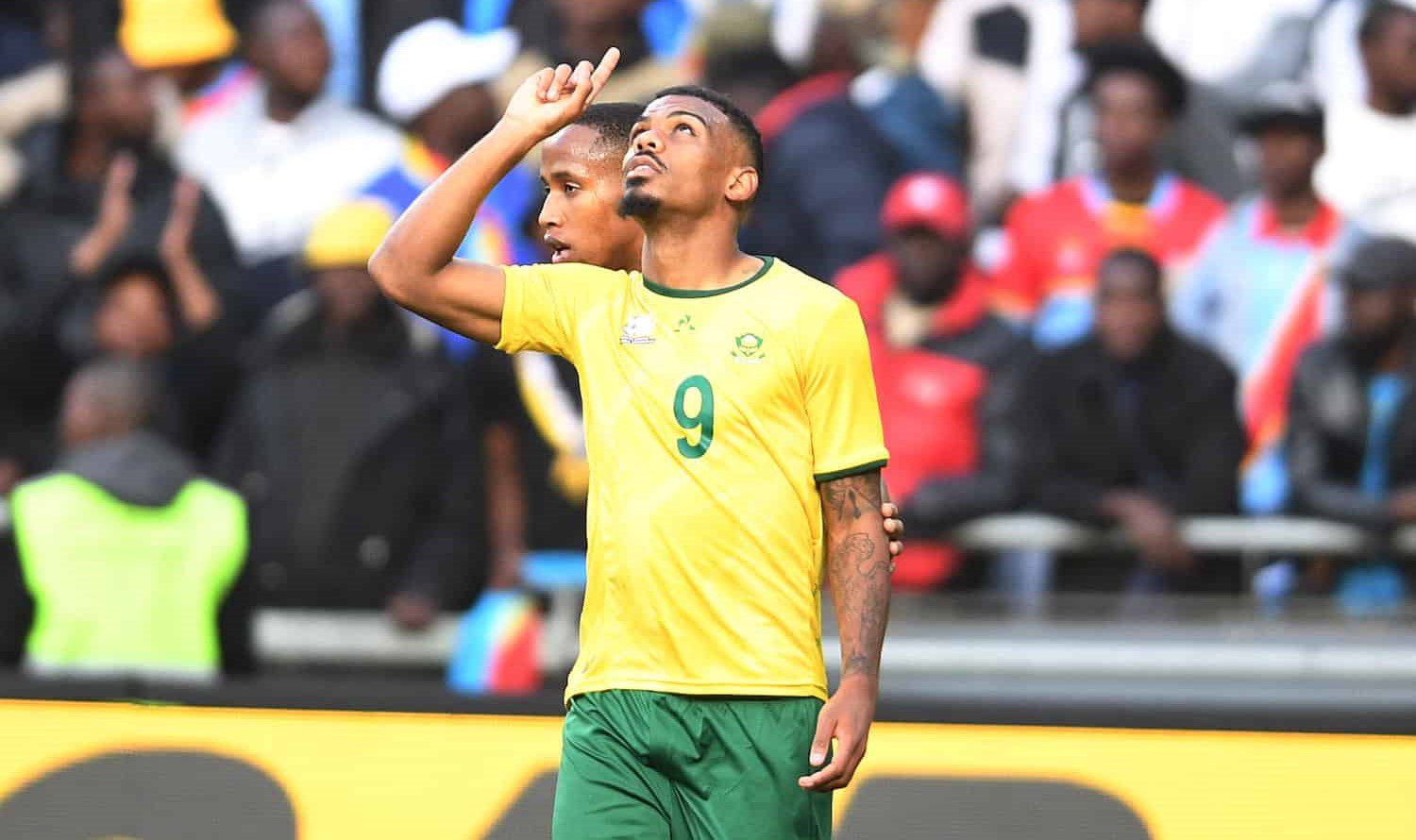 South African footballer Lyle Foster wearing the number 9 yellow and green Bafana Bafana national jersey, pointing upward during a match with spectators in the background. Image used for Sportxparte news. 