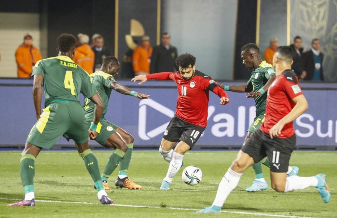 Egyptian forward Mohamed Salah (number 10 in a red jersey) controlling the ball and driving past green-shirted Senegalese defenders, including Pape Abou Cissé (number 4). Another Egyptian player (number 11) runs alongside him to the right in support. Image used for Sportxparte news. 