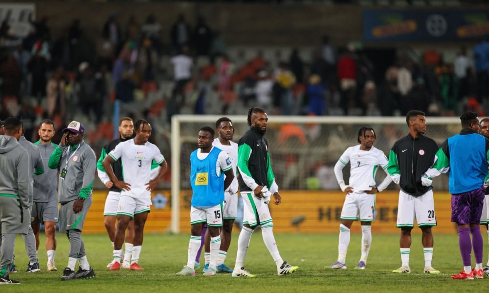 A group of players and staff from the Nigeria men's national football team, the Super Eagles, stand on a stadium pitch looking dejected or pensive. Image used for Sportxparte news.