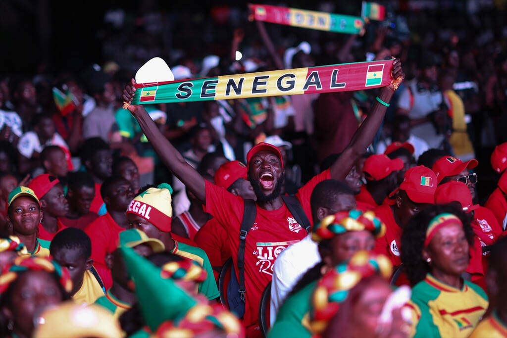 Senegalese soccer fans are cheering passionately as they watch a live broadcast of their national team at a public viewing event. The central supporter is holding up a green, yellow, and red scarf emblazoned with the word "SENEGAL" and the national flag. Image used for Sportxparte news.