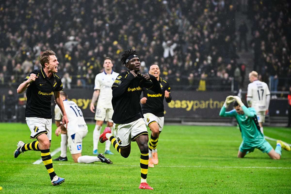 In the frame, Zadok Yohanna (center) wheels away in celebration wearing AIK's iconic black and yellow hooped kit, accompanied by teammate Johan Hove running alongside him on the left. On the right, the devastated Halmstads BK goalkeeper is shown on his knees. Image used for Sportxparte news.