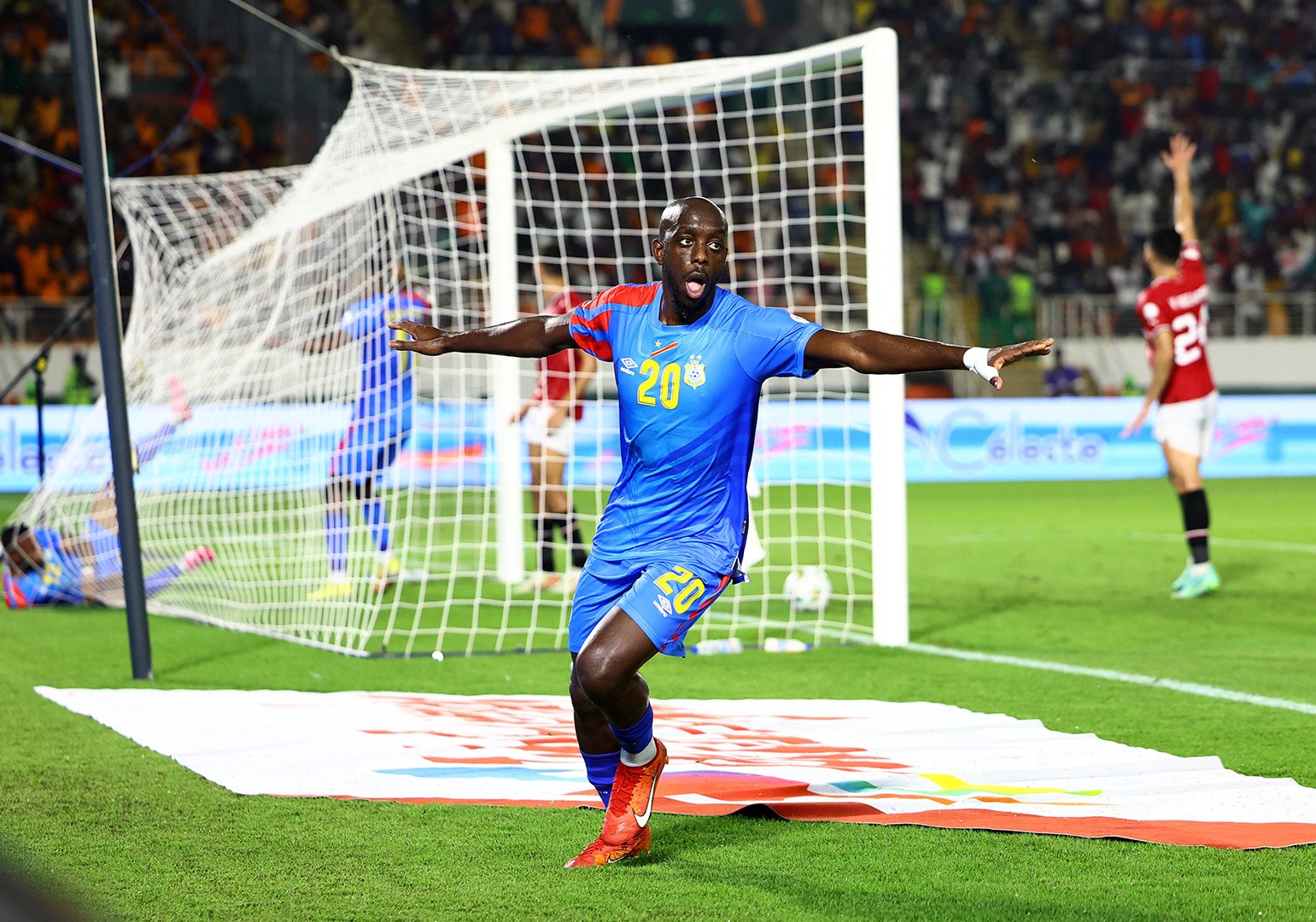 DR Congo professional footballer Yoane Wissa, wearing a blue jersey with number 20, celebrates on the pitch with outstretched arms after a match against Egypt. Image used for Sportxparte news. 
