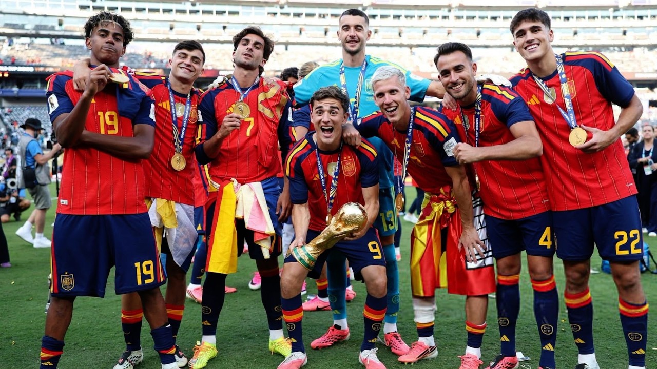 The players, wearing their red and blue home kits with gold winner's medals around their necks, are smiling and posing together. In the center, Gavi holds the gold FIFA World Cup trophy high, flanked by teammates including Lamine Yamal (#19), Pedri, Ferran Torres (#7), Arnau Tenas, Dani Olmo, Eric García, and Pau Cubarsí (#4) in a crowded stadium environment. Image used for Sportxparte news.