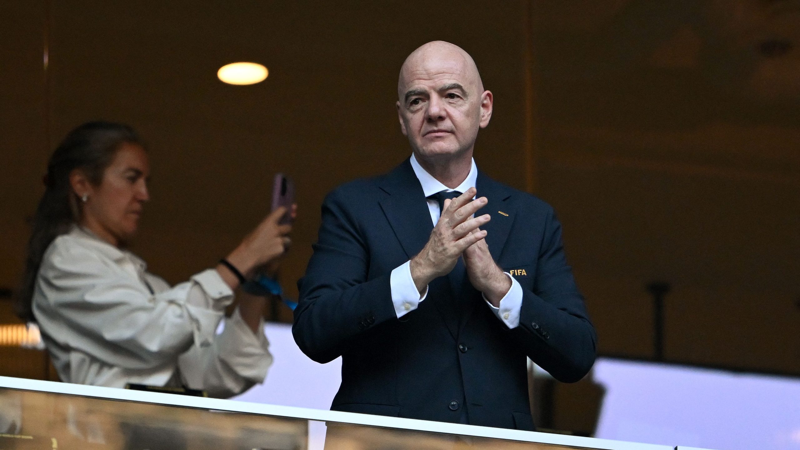 Gianni Infantino, the President of FIFA, wearing a dark navy suit and white shirt while clapping his hands from a stadium suite during a football match. Image used for Sportxparte news.