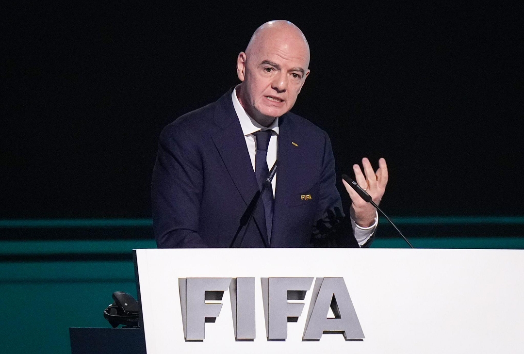 FIFA President Gianni Infantino wearing a dark blue suit and tie, gesturing with his hand while speaking behind a white podium featuring the FIFA logo. Image used for Sportxparte news.