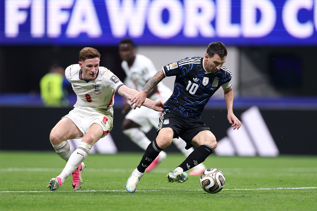 Lionel Messi in a blue Argentina jersey dribbling a soccer ball past Elliot Anderson in a white England jersey during a match. Image used for Sportxparte news.
