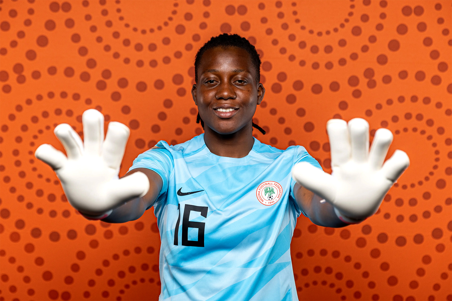 Nigeria women's national football team goalkeeper Chiamaka Nnadozie smiling in a light blue jersey with the number 16 and holding up white goalkeeper gloves against an orange dotted background. Image used for Sportxparte news.