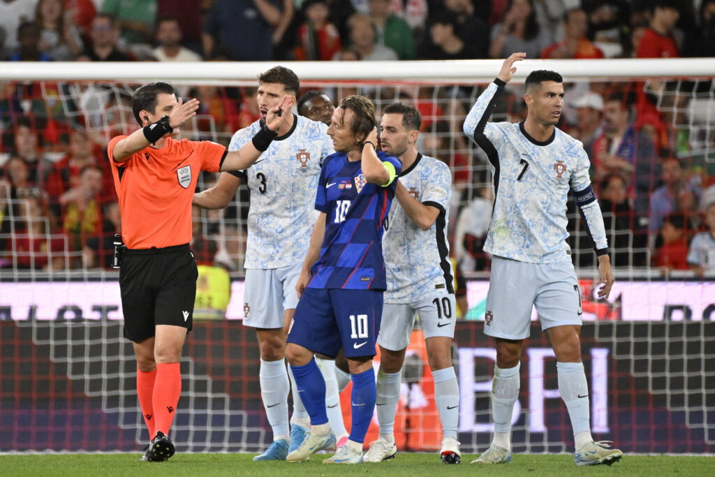 Luka Modrić looks frustrated next to Cristiano Ronaldo and a referee during a Portugal vs Croatia football match. Image used for Sportxparte news.